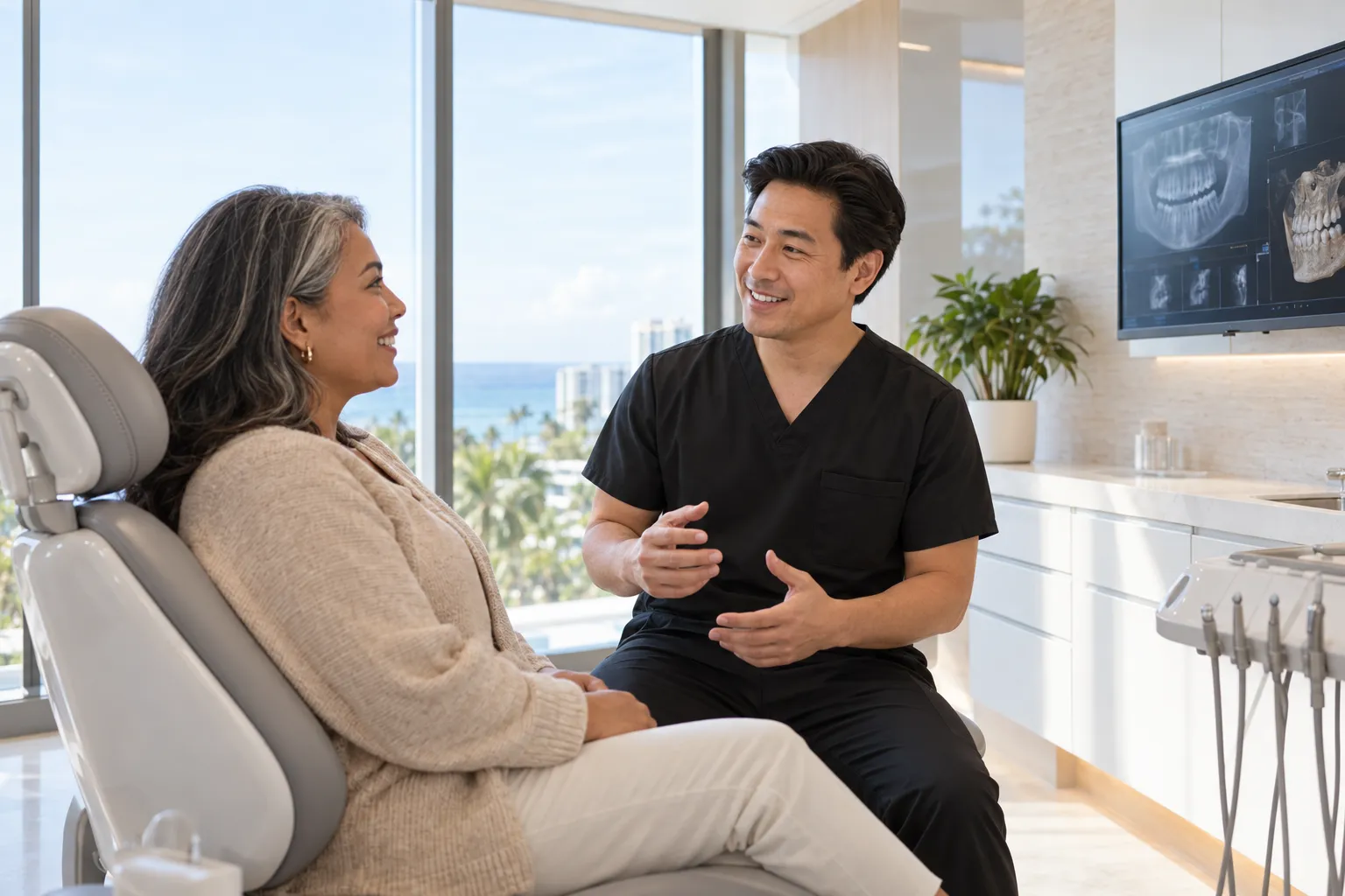 A dentist in black scrubs talks with a patient seated in a bright exam room.