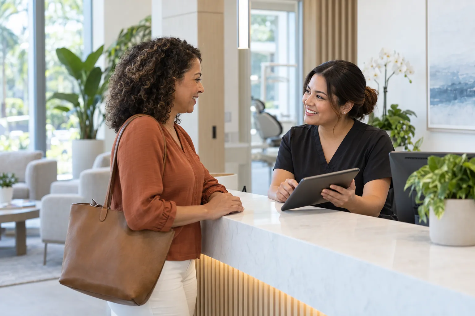 A dental receptionist holding a tablet speaks with a woman at a bright clinic front desk.