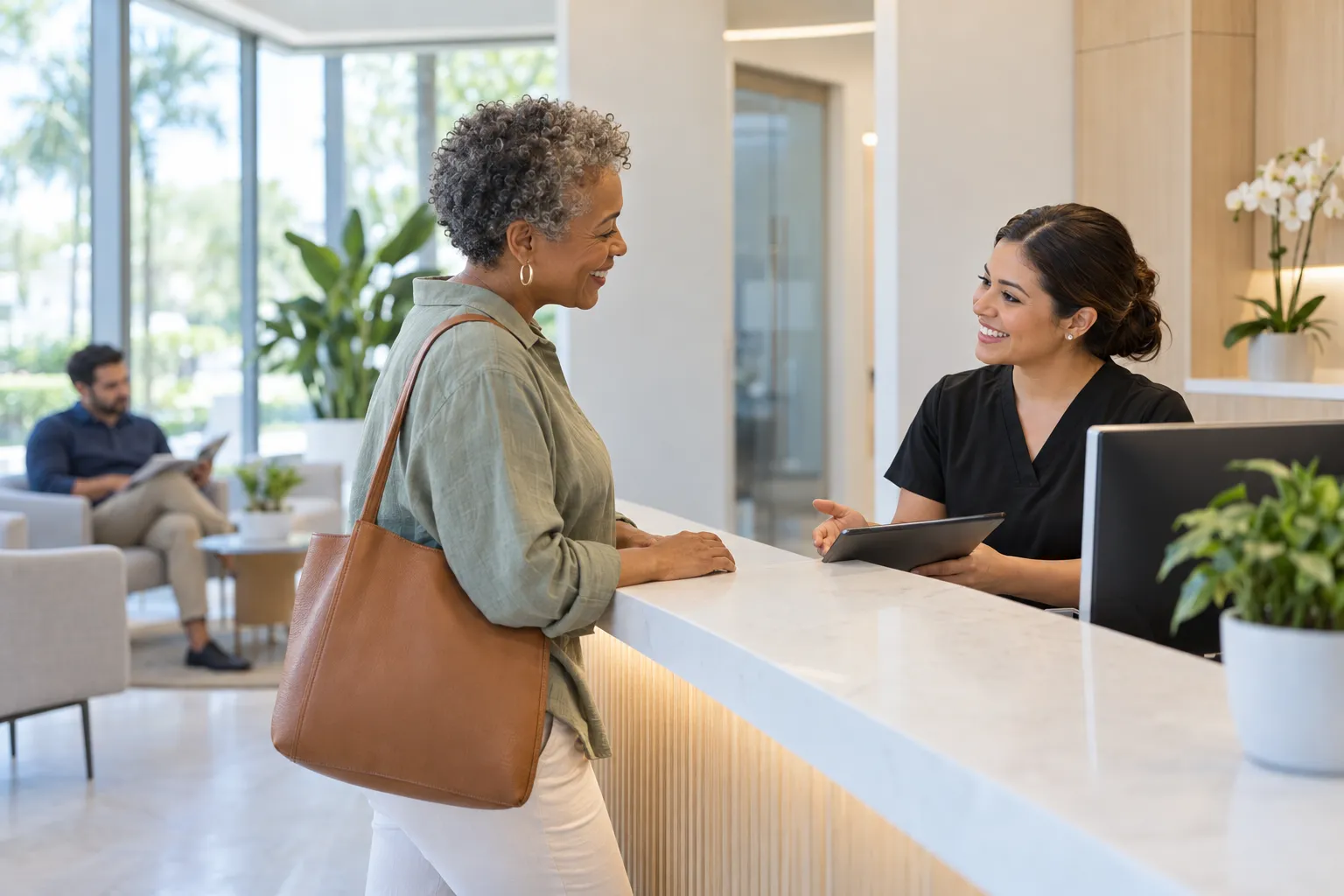 An older woman speaks with a receptionist holding a tablet at a bright dental office front desk.