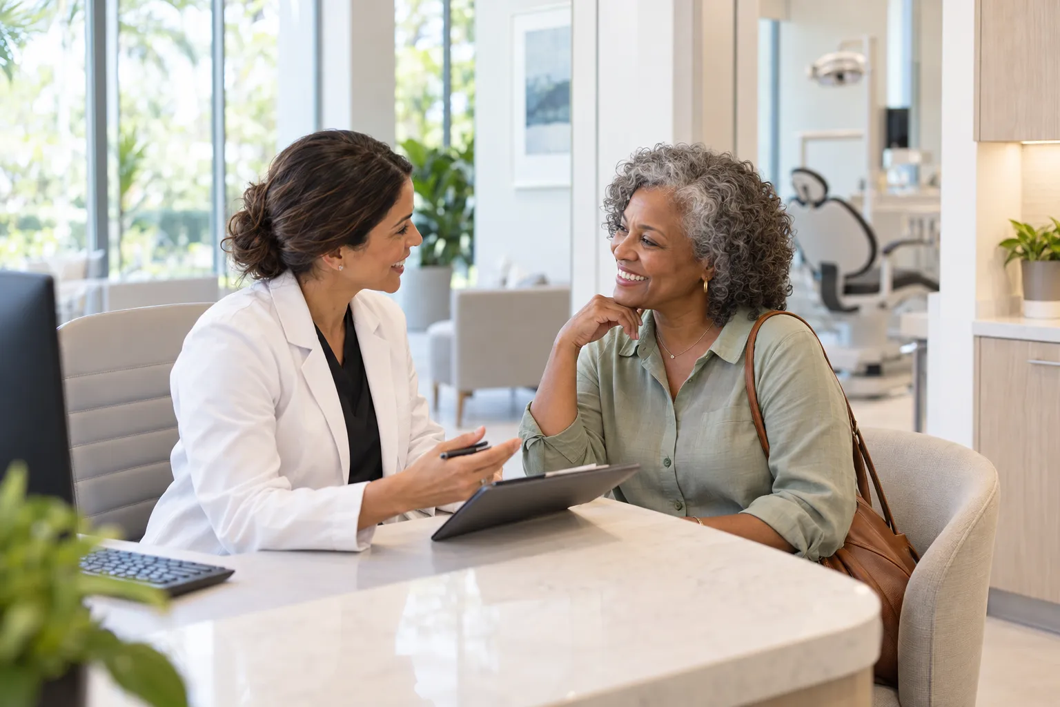 A dentist in a white coat speaks with a smiling patient across a desk in a bright dental office.